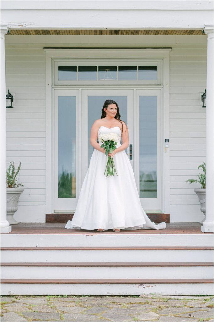 Bride stands on porch in bridal gown for Maine Barn Wedding